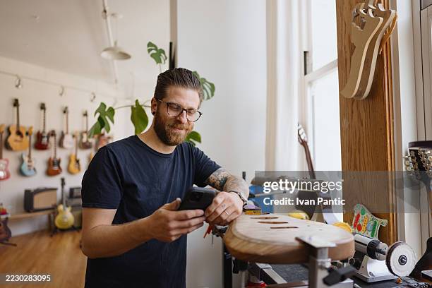 luthier vérifie son smartphone dans son atelier, faisant une pause dans la construction d’un corps de guitare - représentation masculine photos et images de collection