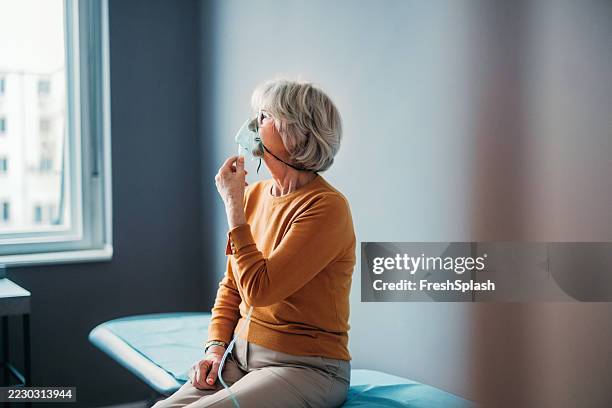 elderly woman using oxygen mask while looking out the window in a clinic - medical oxygen equipment stock pictures, royalty-free photos & images