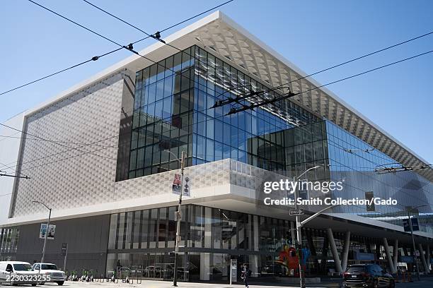 Modern glass facade of Moscone Center expansion with street traffic, overhead trolley wires, and e-scooters under clear sky, San Francisco,...