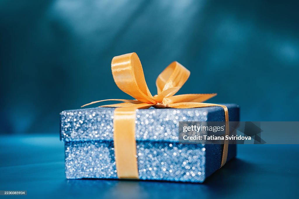Blue gift box with yellow bow ribbon on blue gradient background. Slightly defocused with glitter bokeh