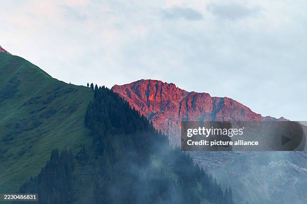 July 2025, Austria, Berwang: Landscape photograph of the red glowing rugged rock face in front of the Suwaldspitze peaks in the reddish light of the...
