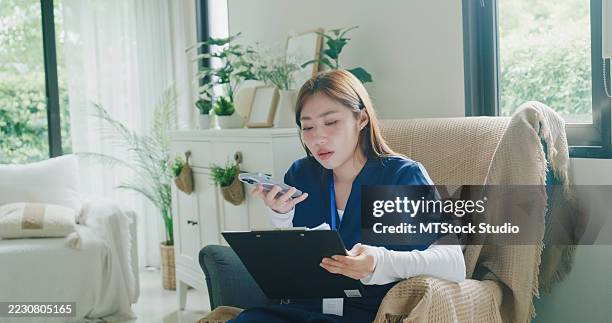 junge asiatische krankenschwester in blauen kitteln nutzt spracherkennungstechnologie am telefon, während sie die patientenversorgungsdokumente auf einem klemmbrett im wohnzimmer zu hause überprüft. krankenschwester nachhaltiges lifestyle-konzept. - medizinische dokumente stock-fotos und bilder