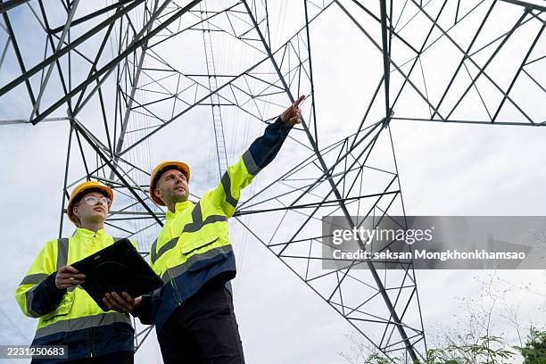 male and female electrical engineers discussing while using technologies under electricity pylon at power station. professional electricians work on laptop while standing near power lines - telephone lines stock pictures, royalty-free photos & images