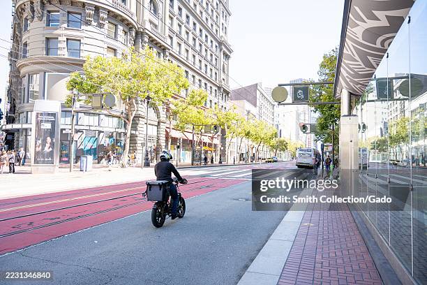 Motorcyclist in delivery gear travels along red transit lane past historic flatiron building and glass bus shelter on Market Street, San Francisco,...