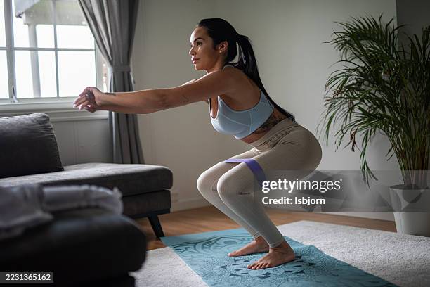 vista lateral de una mujer deportiva haciendo sentadillas con una mini banda de goma en la sala de estar. - goma elástica fotografías e imágenes de stock
