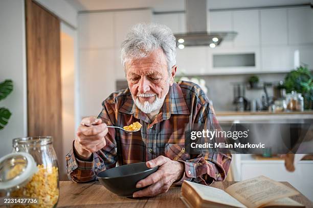 älterer mann genießt eine schüssel müsli und liest in seiner küche - einzelner senior stock-fotos und bilder
