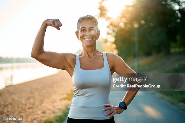 mujer mayor flexionando bíceps después de hacer ejercicio en el parque - flexionar los músculos fotografías e imágenes de stock