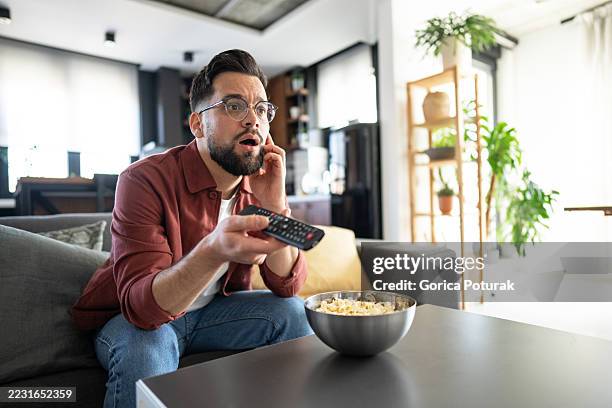 shocked man watching tv and talking on phone with popcorn - man remote control stock pictures, royalty-free photos & images
