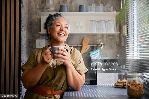 femme mûre heureuse buvant du café dans la cuisine et détournant le regard - baby boomer photos et images de collection