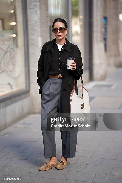 Patricia Wirschke is seen wearing a brown trench coat with buttons and collar; a brown cardigan buttoned up over a white t-shirt with round collar; a...