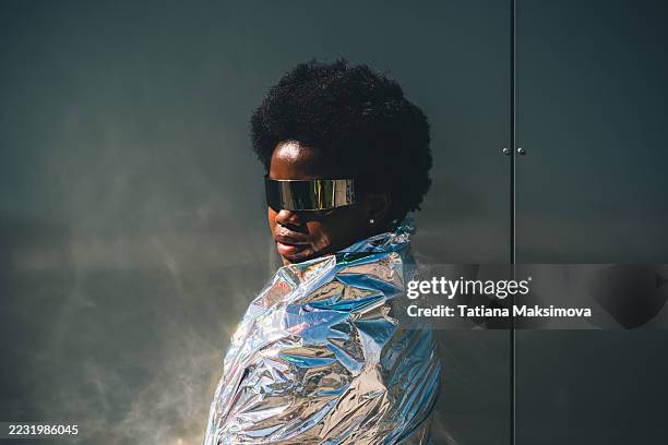 a futuristic portrait of a smiling african american woman wearing reflective visor sunglasses and wrapped in a metallic cloak. - vanguardista imagens e fotografias de stock