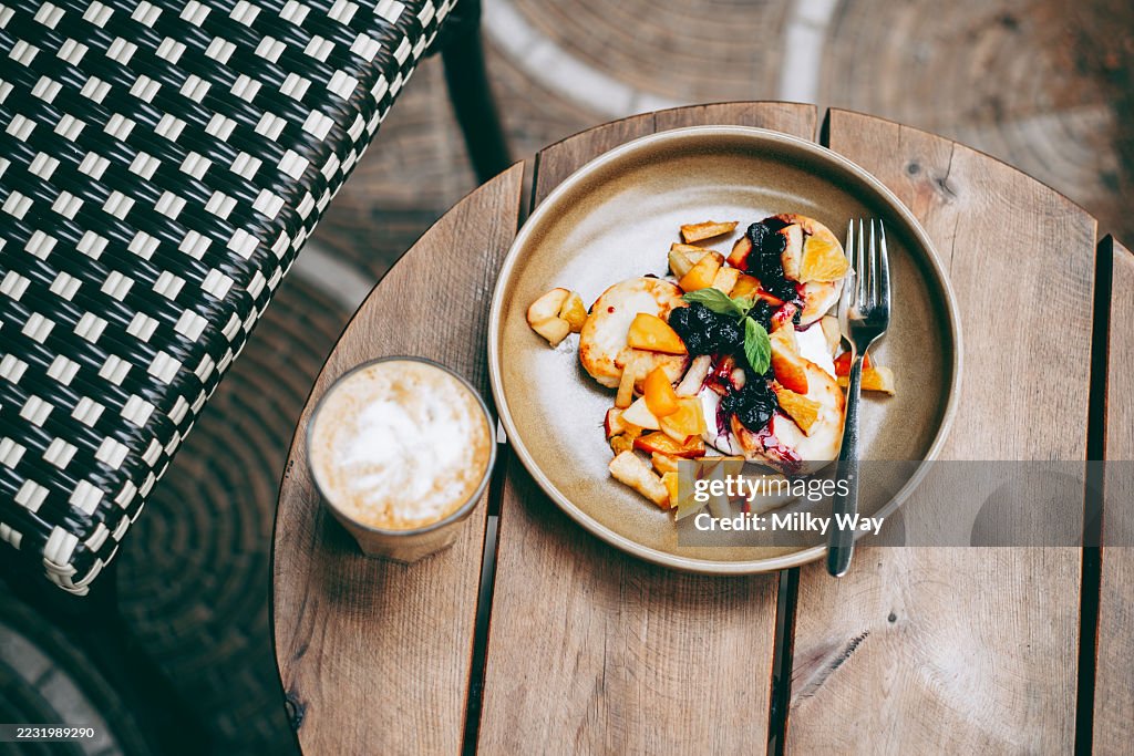 Plate of cottage cheese pancakes or ricotta cheesecakes topped with berries, peach slices, and syrup next to a glass of latte. Breakfast, dessert and coffee break concept.