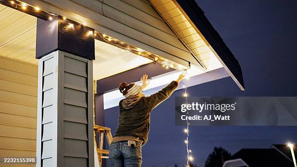 woman attaching christmas lights on house exterior for holiday celebration - hanging stock pictures, royalty-free photos & images
