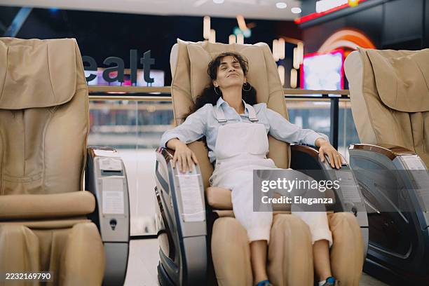 entspannte frau, die in einem massagesessel sitzt und nach dem einkaufen in einem großen einkaufszentrum einen moment der ruhe genießt. sie wirkt ruhig und zufrieden und macht eine pause von einem anstrengenden shopping-therapietag. - massagestuhl stock-fotos und bilder