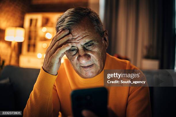 senior man feeling stressed at home in the evening while reading something on his smartphone - low lighting stock pictures, royalty-free photos & images