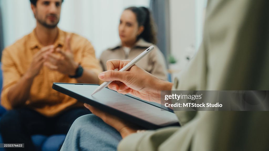 Closeup of young couple in serious discussion with mental health professional during marriage counseling session sit on sofa in living room at home. Therapy session mental health concept.