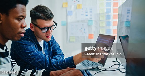 Two male software developers meeting on multiple computer screens displaying programming code, collaborating on project in office. Artificial intelligence and programming. Two male software developers meeting on multiple computer screens displaying programming code, collaborating on project in office. Artificial intelligence and programming.