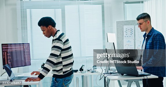 Two male software developers analyzing data coding and working on multiple screens standing at desk in office. Artificial intelligence and programming. Two male software developers analyzing data coding and working on multiple screens standing at desk in office. Artificial intelligence and programming.