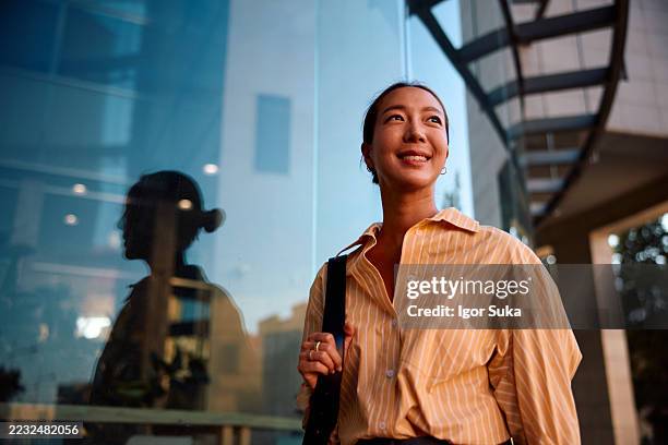 young asian businesswoman smiling and looking up near modern office building - anticipation stock pictures, royalty-free photos & images