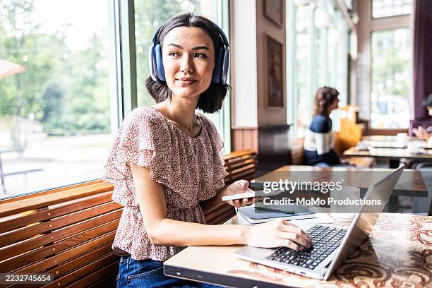 young woman wearing headphones and working on laptop in coffeeshop - freelance work stock pictures, royalty-free photos & images