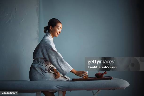 woman in robe enjoying tea while relaxing in minimalist setting - traditional ceremony stock pictures, royalty-free photos & images
