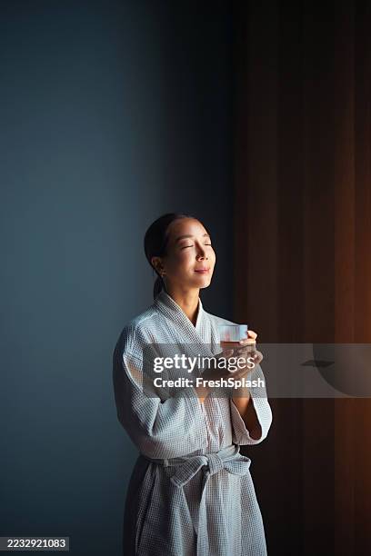 tranquil woman enjoying a warm beverage in morning light at home - bathrobe stock pictures, royalty-free photos & images