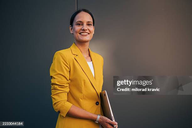 smiling businesswoman holding laptop standing against gray wall - vestido amarelo imagens e fotografias de stock