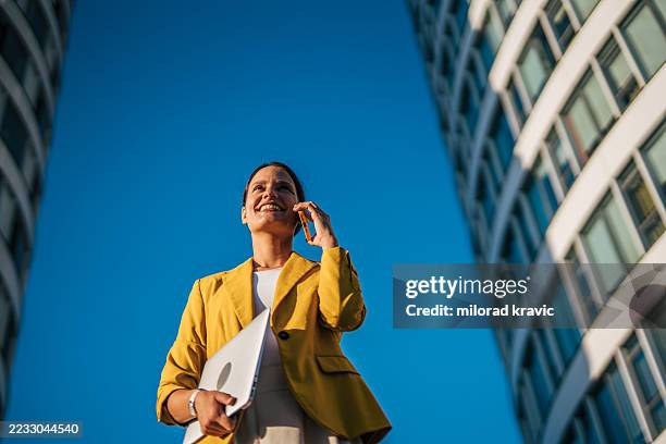 businesswoman talking on phone holding laptop in urban setting - vestido amarelo imagens e fotografias de stock