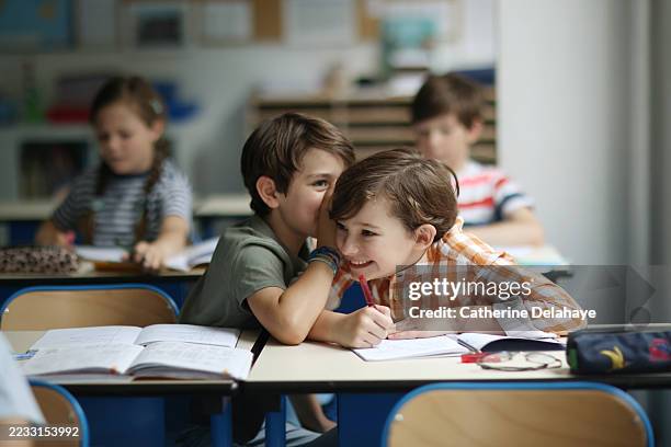 a child whispering in his friend's ear in a classroom - écolier photos et images de collection