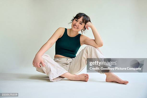 ganzkörperansicht einer frau der generation z, die lässig auf einem studioboden sitzt und sich mit einem verspielten, entspannten lächeln entspannt anlehnt - lehnend stock-fotos und bilder