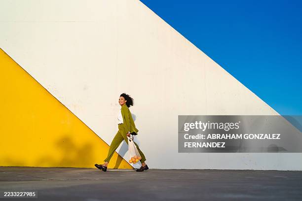 mujer segura de sí misma caminando por la calle urbana, sosteniendo una bolsa de compras ecológica junto a un colorido fondo de pared - vida sencilla fotografías e imágenes de stock