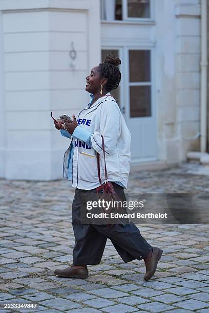 Guest wears braided hair styled in a high bun, red-framed rectangular sunglasses, a gold-toned drop earring and silver rings, a burgundy leather...