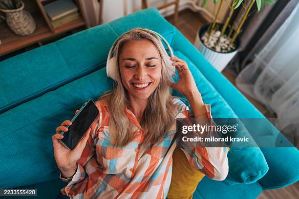 happy woman listening to music on headphones and smartphone while relaxing on sofa - muziekstreamingdienst stockfoto's en -beelden