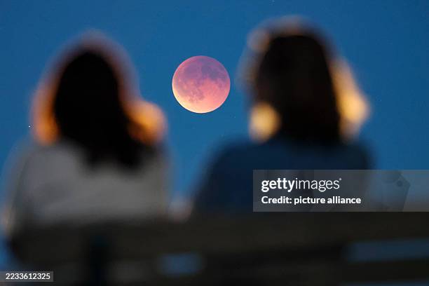 September 2025, Saxony, Leipzig: Onlookers watch the total lunar eclipse, also known as the blood moon, from a park bench. The moon gradually moves...