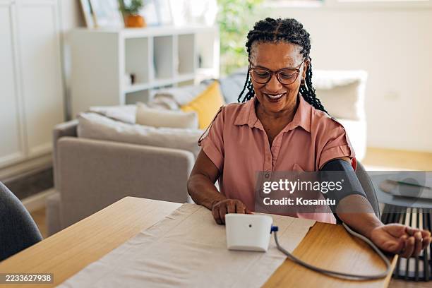 mature woman measures her blood pressure while seated at the dining table in a bright, tidy home. focused and relaxed, she uses a digital device to track her heart health - blood pressure gauge stock pictures, royalty-free photos & images