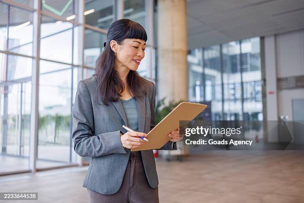 asian mid adult businesswoman writing on clipboard in office lobby - checklist stock pictures, royalty-free photos & images