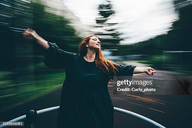 a woman with red hair is spinning on merry go round carousel with her arms outstretched. - playground roundabout stock pictures, royalty-free photos & images