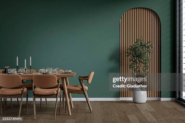 a modern but retro dining room with a full (set) table and chairs in front of a pine green wall and a potted plant in front of a wooden arched alcove, near a window, '70s retro style furniture - parket stockfoto's en -beelden
