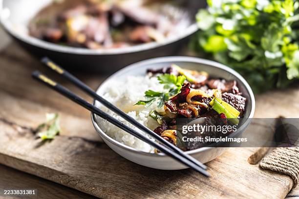 a close-up of a bowl of asian food placed on a wooden board. the bowl contains white rice and stir-fried beef with vegetables such as green onions and caramelized onions, garnished with fresh parsley. - wok stockfoto's en -beelden