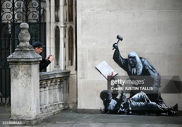 Person takes a photograph of an artwork by street artist Banksy, depicting a judge using a gavel to beat a protester using a placard as protection,...
