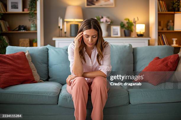 sad woman sitting on sofa touching temples suffering from headache - infertility stock pictures, royalty-free photos & images