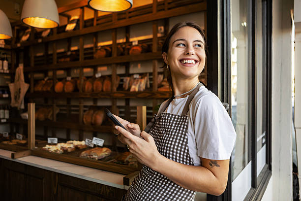 Young adult smiling and holding a phone while looking out of a doorway in her bakery.