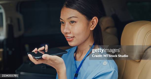 young asian female nurse in blue scrubs sitting in a car, using a voice command feature on her phone. nurse sustainable lifestyle concept. - paramedic phone stock pictures, royalty-free photos & images