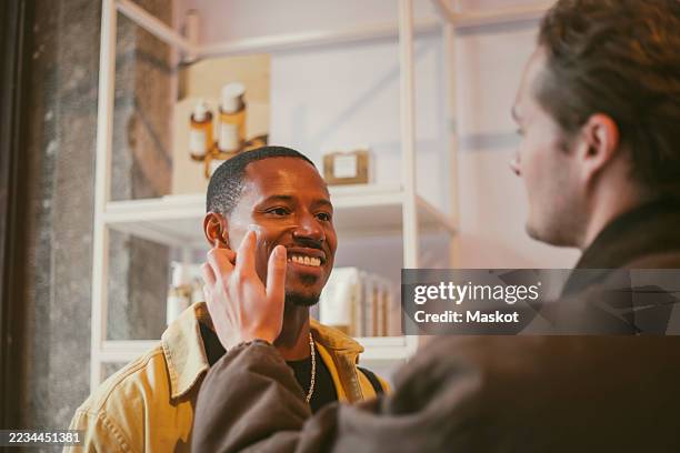 young man applying face cream to smiling male friend while shopping at beauty store - bewogen beeldtechniek stockfoto's en -beelden