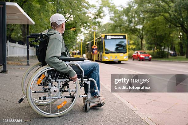 behinderter mann sitzt im rollstuhl und wartet an der bushaltestelle - öffentliches verkehrsmittel stock-fotos und bilder