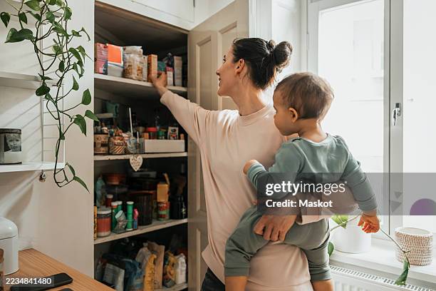 mother carrying baby boy removing ingredient from pantry in kitchen at home - speisekammer stock-fotos und bilder