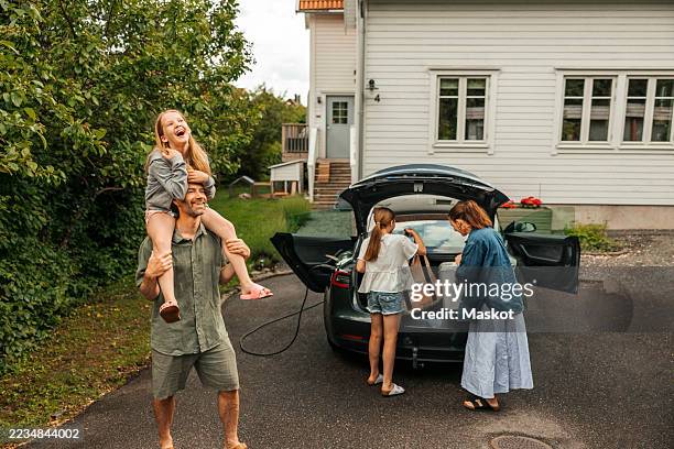 smiling father carrying daughter on shoulder while woman loading car trunk with girl on driveway - piggyback stock pictures, royalty-free photos & images