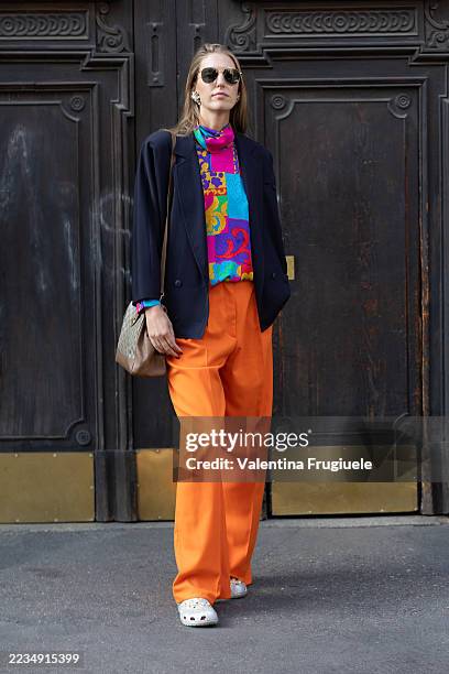 Person wears black sunglasses sunglasses, silver crocs, a multicolor printed turtle neck top, a blue jacket, a beige leather bag, silver earrings and...