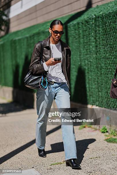 Guest wears a sleek center-parted low bun, narrow black oval sunglasses, a silver ring, a large black crinkled-leather shoulder bag with a wide strap...