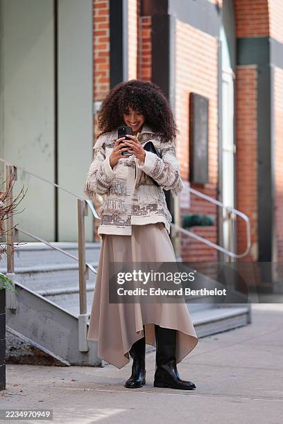 Guest wears shoulder-length curly hair, carrying a structured black leather envelope clutch, a cream and beige boucle jacket with a wide collar,...